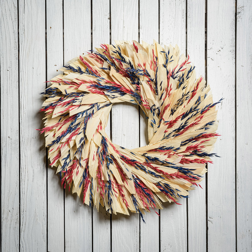 22" wreath made of red and blue oats and natural corn husk on a white wood fence background.