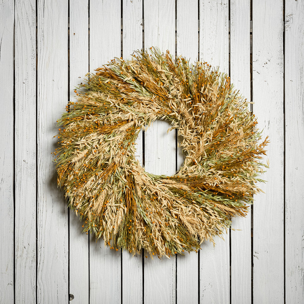 A wreath made of Sunkissed Aveena-oats, dijon Sudan grass, saffron flax, and natural wheat on a white wood background.