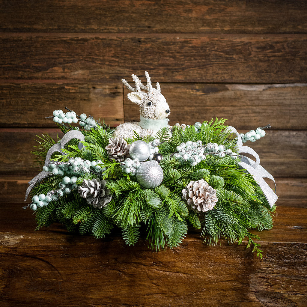 A holiday centerpiece of noble fir, white pine, incense cedar, 2 silver and white ball clusters, 4 light teal-blue glitter berry branches, 2 grey glitter bows, 6 white glitter pinecones, and a white sisal deer sitting on a small wood table with a wood background.