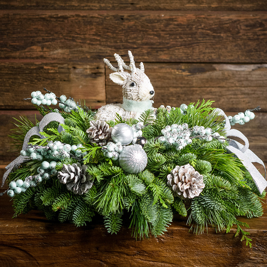 A holiday centerpiece of noble fir, white pine, incense cedar, 2 silver and white ball clusters, 4 light teal-blue glitter berry branches, 2 grey glitter bows, 6 white glitter pinecones, and a white sisal deer sitting on a small wood table with a wood background.