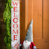 White-washed with a red barn in a wintery holiday snow scene is accented with trees and falling snowflakes with "Welcome" written in large red letters paired with a plush gnome and garland on a front porch.