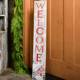 White-washed with a red barn in a wintery holiday snow scene is accented with trees and falling snowflakes with "Welcome" written in large red letters paired with garland on a front porch.