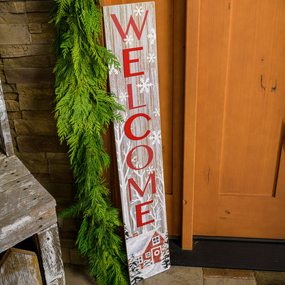 White-washed with a red barn in a wintery holiday snow scene is accented with trees and falling snowflakes with "Welcome" written in large red letters paired with garland on a front porch.