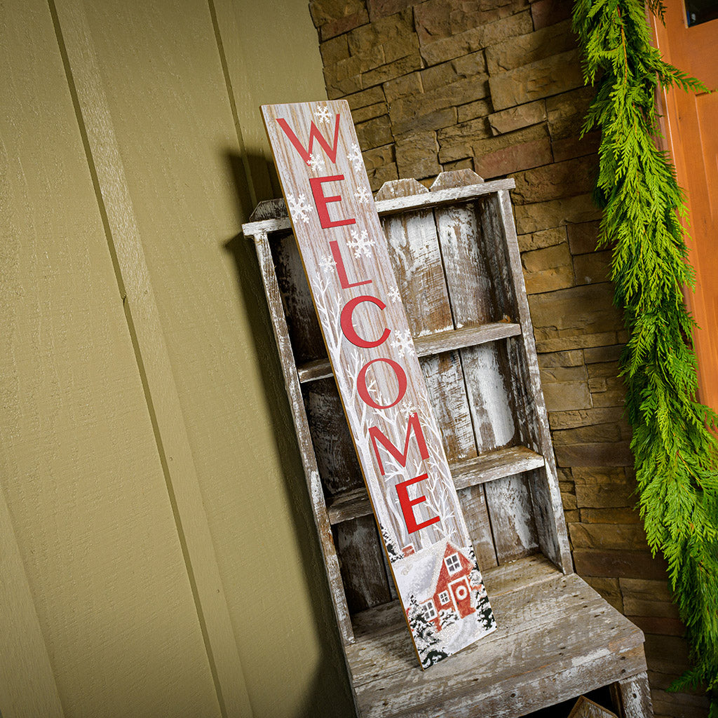 White-washed with a red barn in a wintery holiday snow scene is accented with trees and falling snowflakes with "Welcome" written in large red letters sitting on a wood shelf and paired with garland on a front porch.