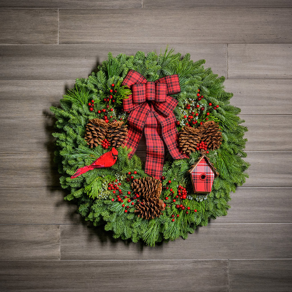 Holiday wreath made of noble fir, white pine, incense cedar, and juniper with ponderosa pine cones, red rosehip/mini pine cone clusters, faux berry clusters, red cardinal, a mini plaid birdhouse, and a red and black plaid bow hanging on a wooden wall