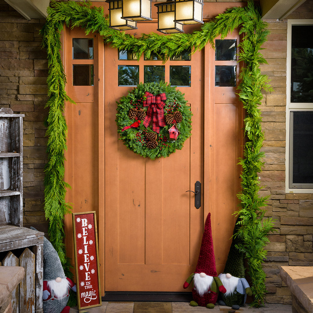 Holiday wreath made of noble fir, white pine, incense cedar, and juniper with ponderosa pine cones, red rosehip/mini pine cone clusters, faux berry clusters, red cardinal, a mini plaid birdhouse, and a red and black plaid bow hanging on a front door