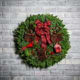 Holiday wreath made of noble fir, white pine, incense cedar, and juniper with ponderosa pine cones, red rosehip/mini pine cone clusters, faux berry clusters, red cardinal, a mini plaid birdhouse, and a red and black plaid bow hanging on a white brick wall