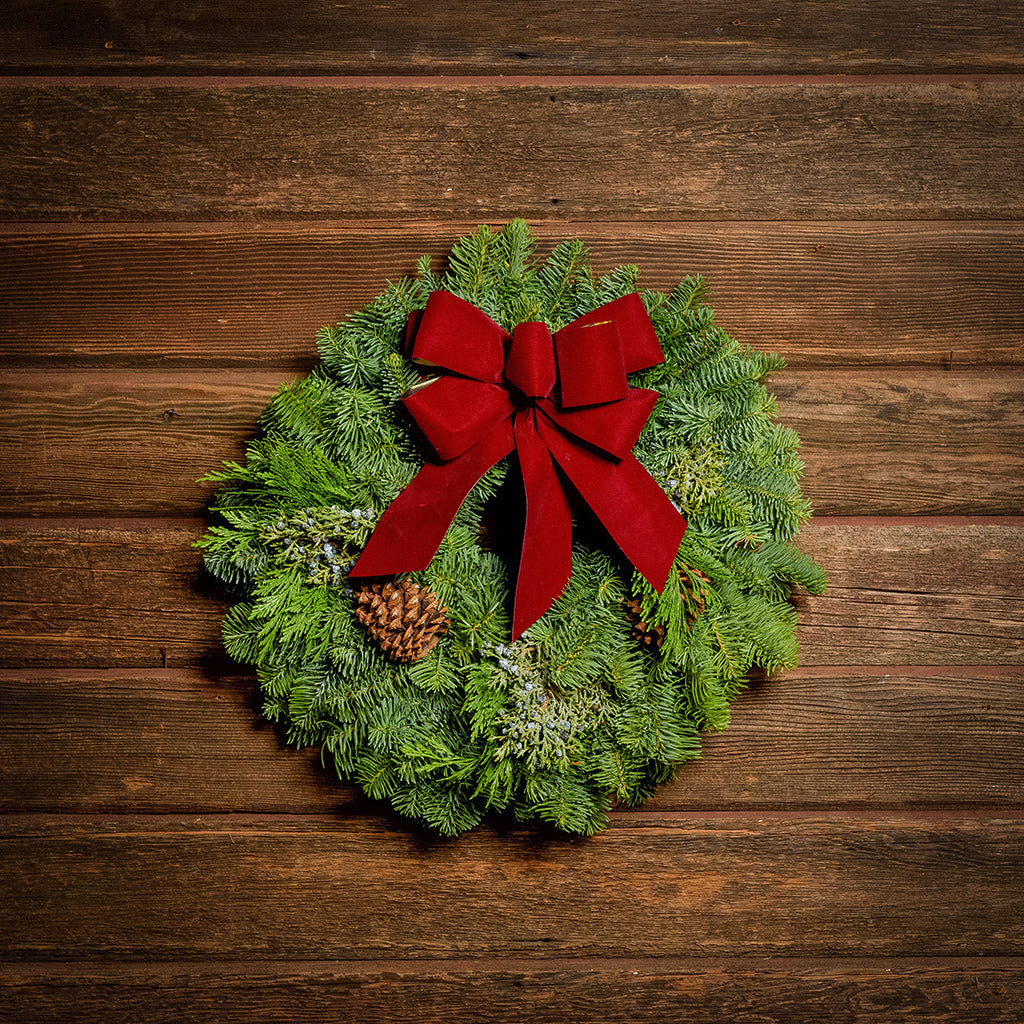 Christmas wreath made of fir, cedar, and juniper with pine cones and a burgundy with gold back bow on a wood background.