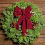 Christmas wreath made of fir, cedar, and juniper with pine cones and a burgundy with gold back bow on a wood background.