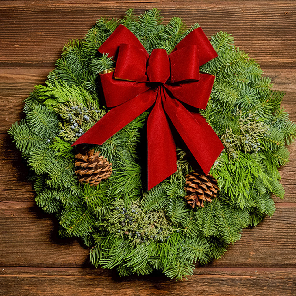 Christmas wreath made of fir, cedar, and juniper with pine cones and a red with gold back bow on a wood background.