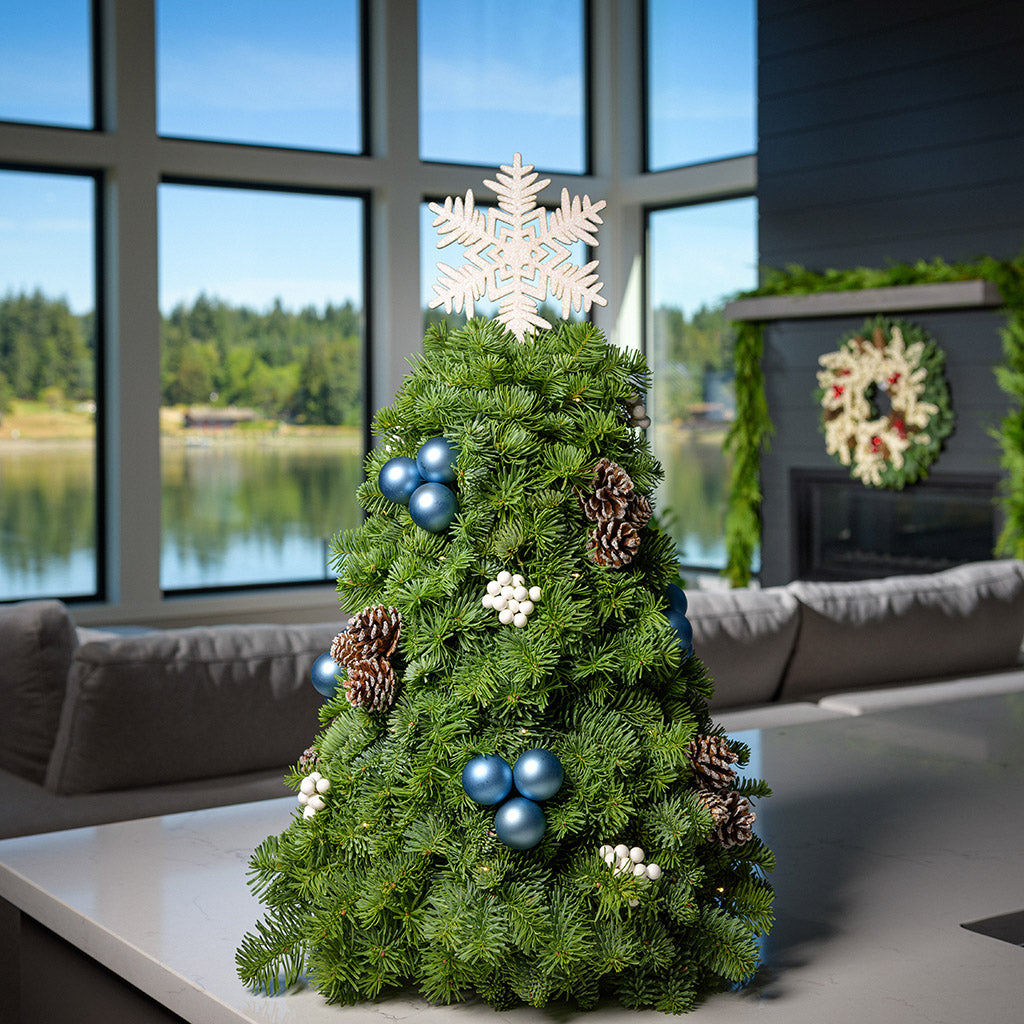 A tree made of noble fir, frosted pinecones, white berry clusters, dark blue ball clusters, a snowflake ornament tree topper, and a strand of warm white lights sitting on a counter.