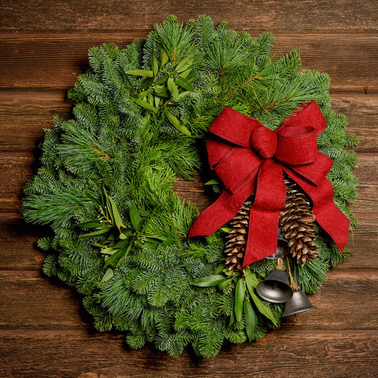 Holiday wreath made of noble fir, incense cedar, white pine, and California bay leaves with 6"- 8" white pine cones, burnished silver bells hanging from a jute string, and a red brushed linen bow on a dark wood background.