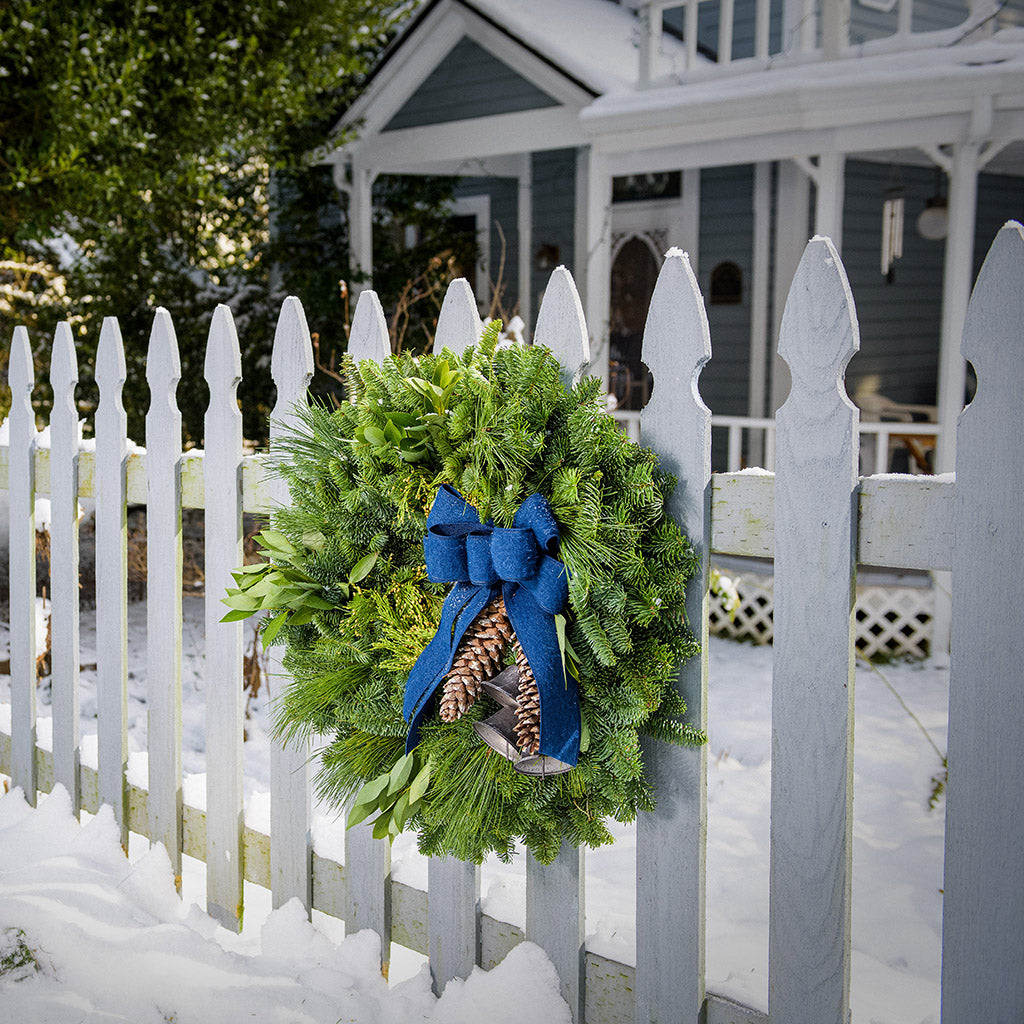 Holiday wreath made of noble fir, incense cedar, white pine, and California bay leaves with 6"- 8" white pine cones, burnished silver bells hanging from a jute string, and a navy blue brushed linen bow hanging on a white picket fence.