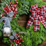 A holiday wreath of noble fir and western cedar with a woodsy grey plush gnome, iced red berry twigs, 6 silver glitter cones, and a grey and red linen flocked buffalo plaid bow on a wood background