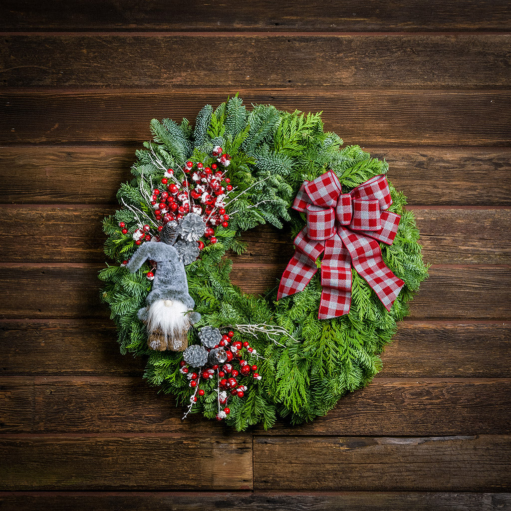 A holiday wreath of noble fir and western cedar with a woodsy grey plush gnome, iced red berry twigs, 6 silver glitter cones, and a grey and red linen flocked buffalo plaid bow on a wood background.