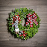 A holiday wreath of noble fir and western cedar with a woodsy grey plush gnome, iced red berry twigs, 6 silver glitter cones, and a grey and red linen flocked buffalo plaid bow on a wood background.