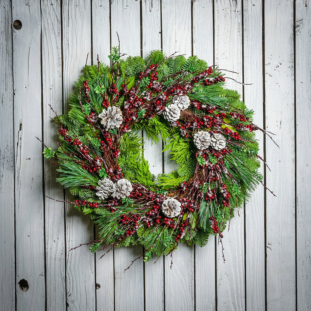 Christmas wreath made of noble fir, cedar, white pine, and bay leaves with 9 frosted Australian pine cones, and a ring of frosted branches with faux burgundy berries hanging on a white fence