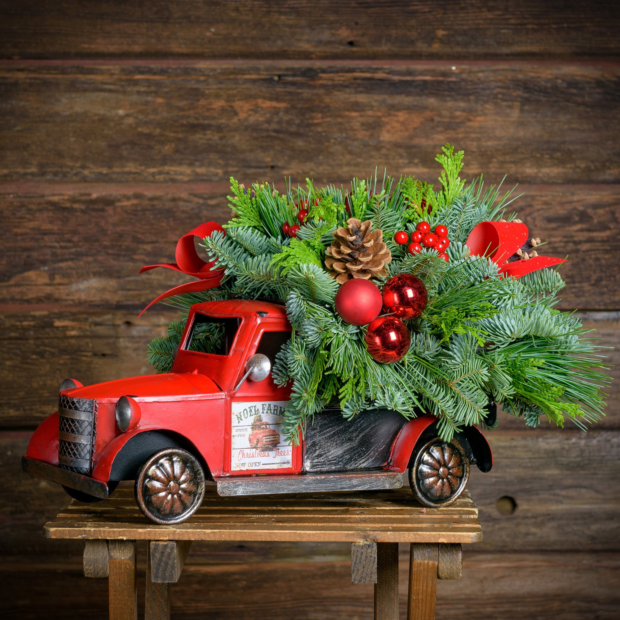 Fresh greens, pine cones,red ball & berry clusters,red velvet bow tucks in a metal truck with removable “Noel Farms” magnets 